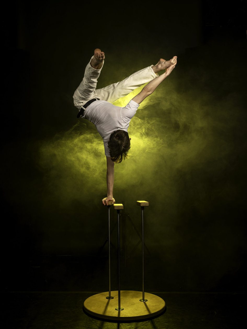 Eine junger Mann steht im Handstand auf einer Hand auf einer Handstandstütze, die Beine in V-Form geöffnet. Gelbes Licht kommt von hinten.