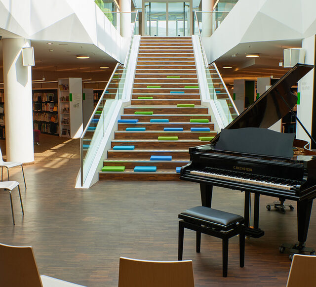 Un piano de cola en la biblioteca musical frente a una escalera y estanterías.