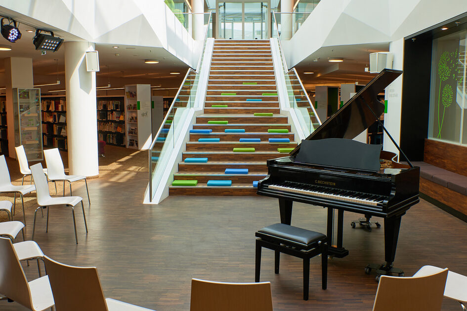 Ein Konzertflügel in der Musikbibliothek vor einer Treppe und Bücherregalen.