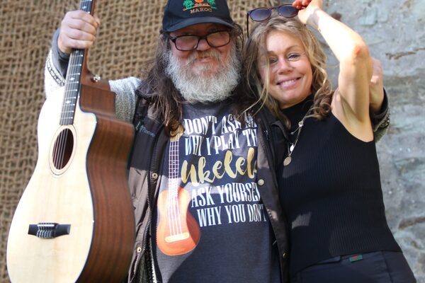 A la izquierda un hombre con barba blanca, gafas y guitarra, a la derecha una mujer de negro con gafas de sol en la cabeza sonriendo. Ambos se abrazan despreocupadamente.