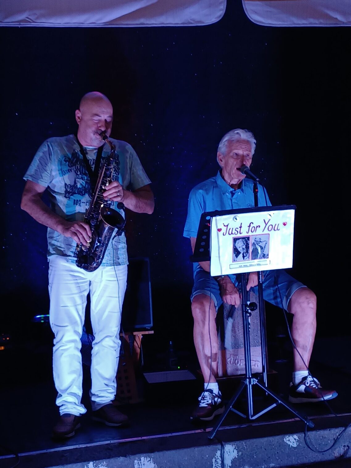 Το δίδυμο Just for you στη σκηνή. Στα αριστερά ένας άνδρας με σαξόφωνο, στα δεξιά ένας άνδρας που κάθεται στο cajon και τραγουδάει στο μικρόφωνο.