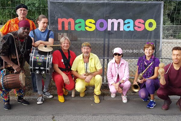 8 people stand with instruments in front of a black banner with colorful lettering "masomaso". Each person is dressed in a different bright color. Just like the clothes, the people are also very different in terms of age and origin.