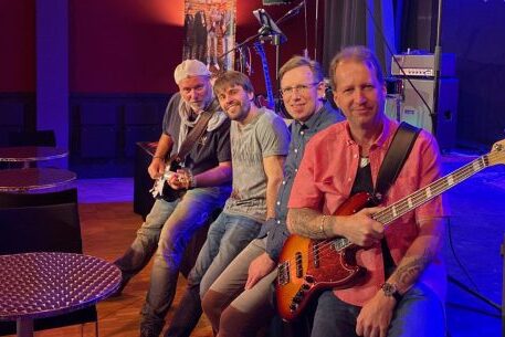 I quattro membri della band sono seduti su un palco, fotografati di lato. Un uomo all'estrema sinistra con una chitarra, entrambi gli uomini al centro non tengono uno strumento, l'uomo all'estrema destra tiene un basso. Sono seduti sul bordo del palco, che è illuminato a colori.