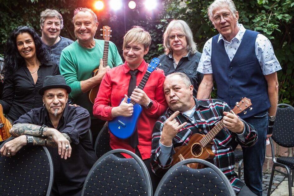 Ομαδική φωτογραφία των Stagies. Υπάρχουν οκτώ άτομα. Οι περισσότεροι είναι ντυμένοι στα μαύρα και δύο στα πράσινα και κόκκινα. Τρεις κρατούν διαφορετικά γιουκαλίλι.  Όλοι φαίνονται χαλαροί.