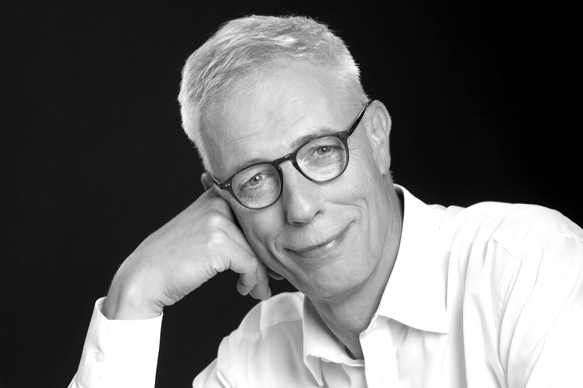 Alexander von Wangenheim sonríe a la cámara. La foto tiene un filtro en blanco y negro. Alexander von Wangenheim apoya la cabeza en la mano.