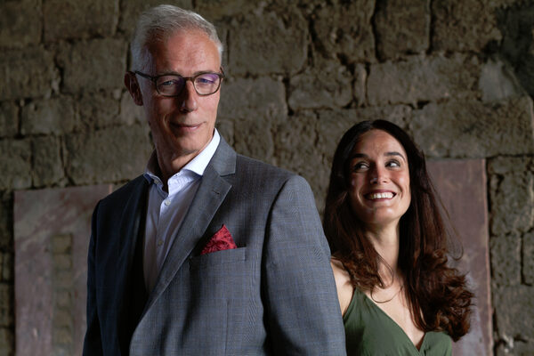 On the right is a man in a suit and to his right is a woman in a green dress. The man looks into the camera and the woman looks up at him and smiles. They are standing in front of an old stone wall.