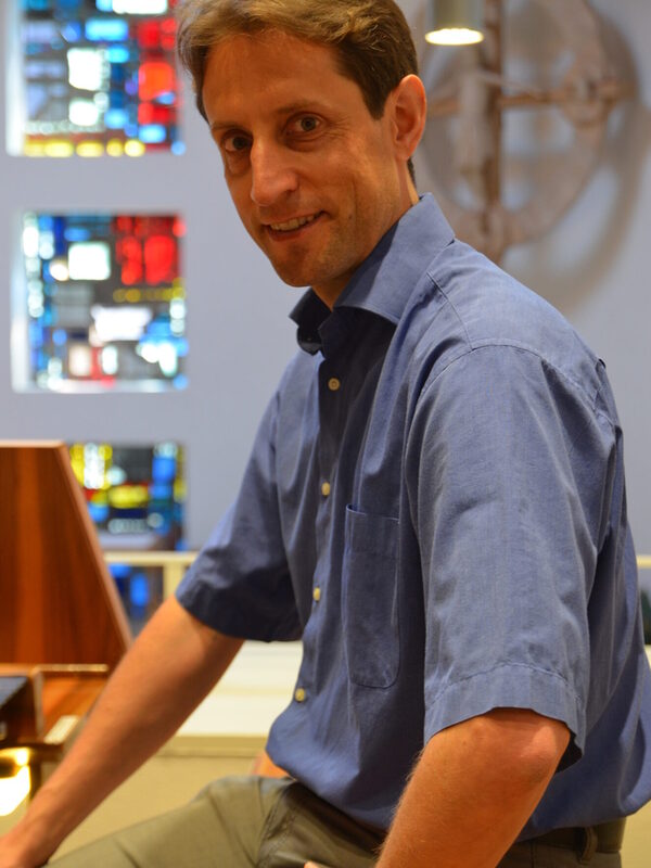 Uomo in camicia blu seduto a un organo. Guarda bene nella telecamera.