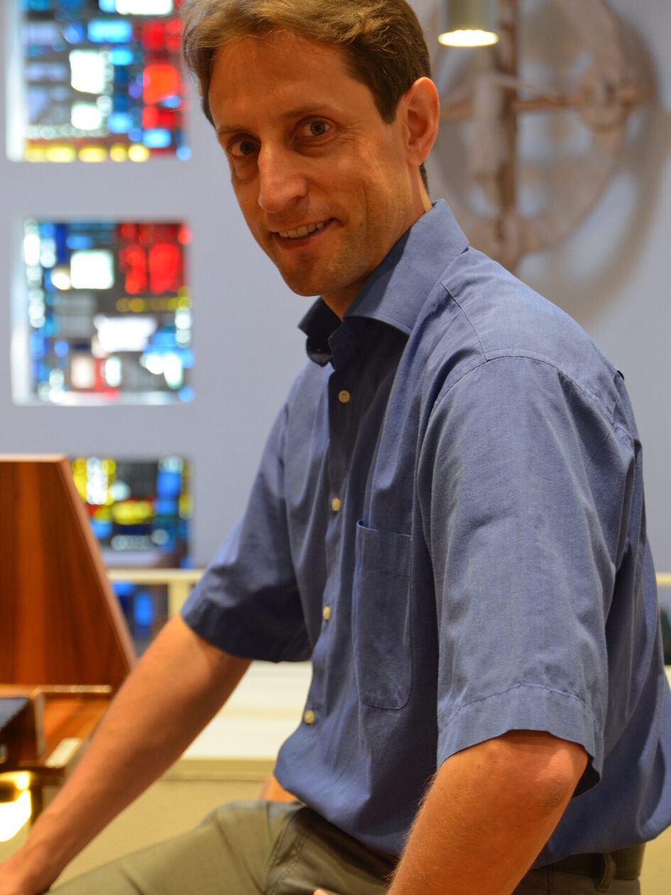 Uomo in camicia blu seduto a un organo. Guarda bene nella telecamera.