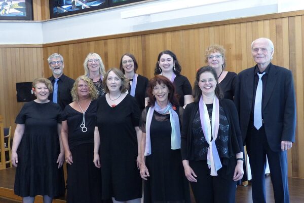Un coro di undici persone si trova davanti a una telecamera. Sono tutti vestiti di nero e in modo elegante e sorridono alla telecamera. Ci sono due uomini e nove donne.