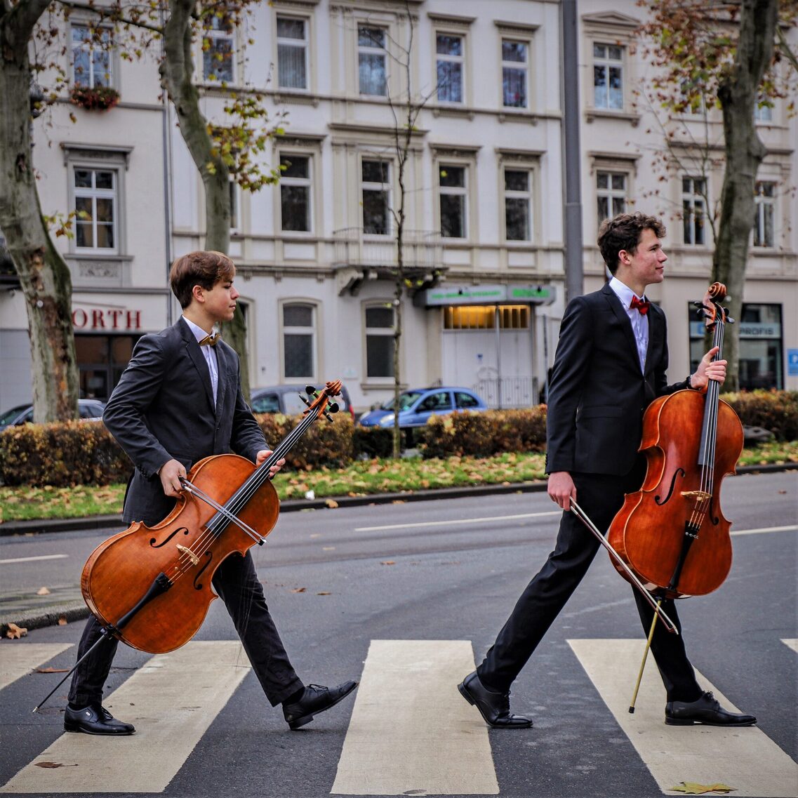 Doi tineri violonceliști, îmbrăcați la costum și cu violoncelul în mână, traversează o alee pietonală. Privesc concentrați în lateral.