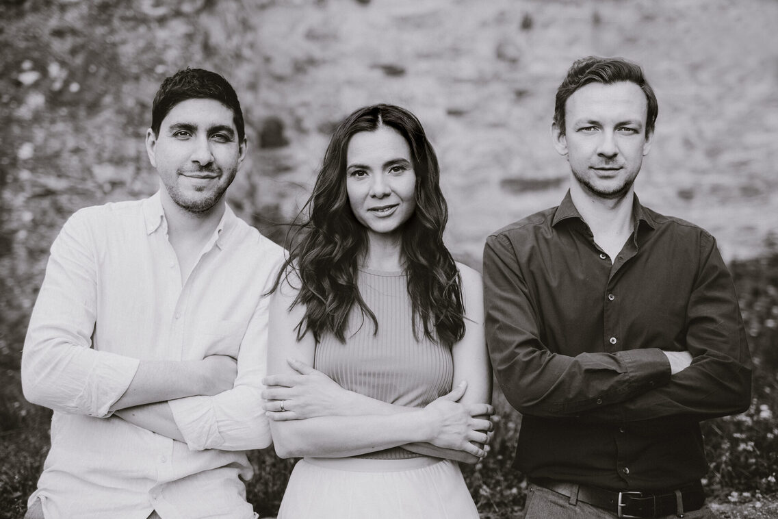 Μια ασπρόμαυρη ομαδική φωτογραφία του τρίο στη φύση. Και οι τρεις έχουν τα χέρια τους σταυρωμένα μπροστά από το πάνω μέρος του σώματός τους και χαμογελούν στην κάμερα.