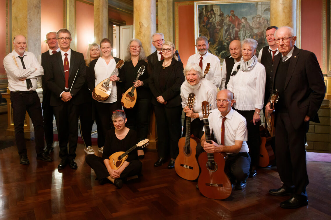 Das Mandolinenorchester steht mit den Instrumenten zusammen. Alle sind in Konzertkleidung und man sieht einen Mann mit einem Dirigentenstab.