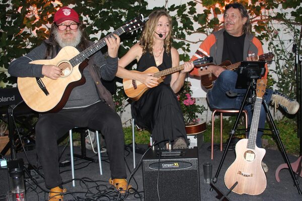 The band is sitting on chairs on a stage. On the left is a sturdy bearded man with a guitar, in the middle a blonde woman in a black dress with a ukulele and microphone, on the right a man with a ukulele.