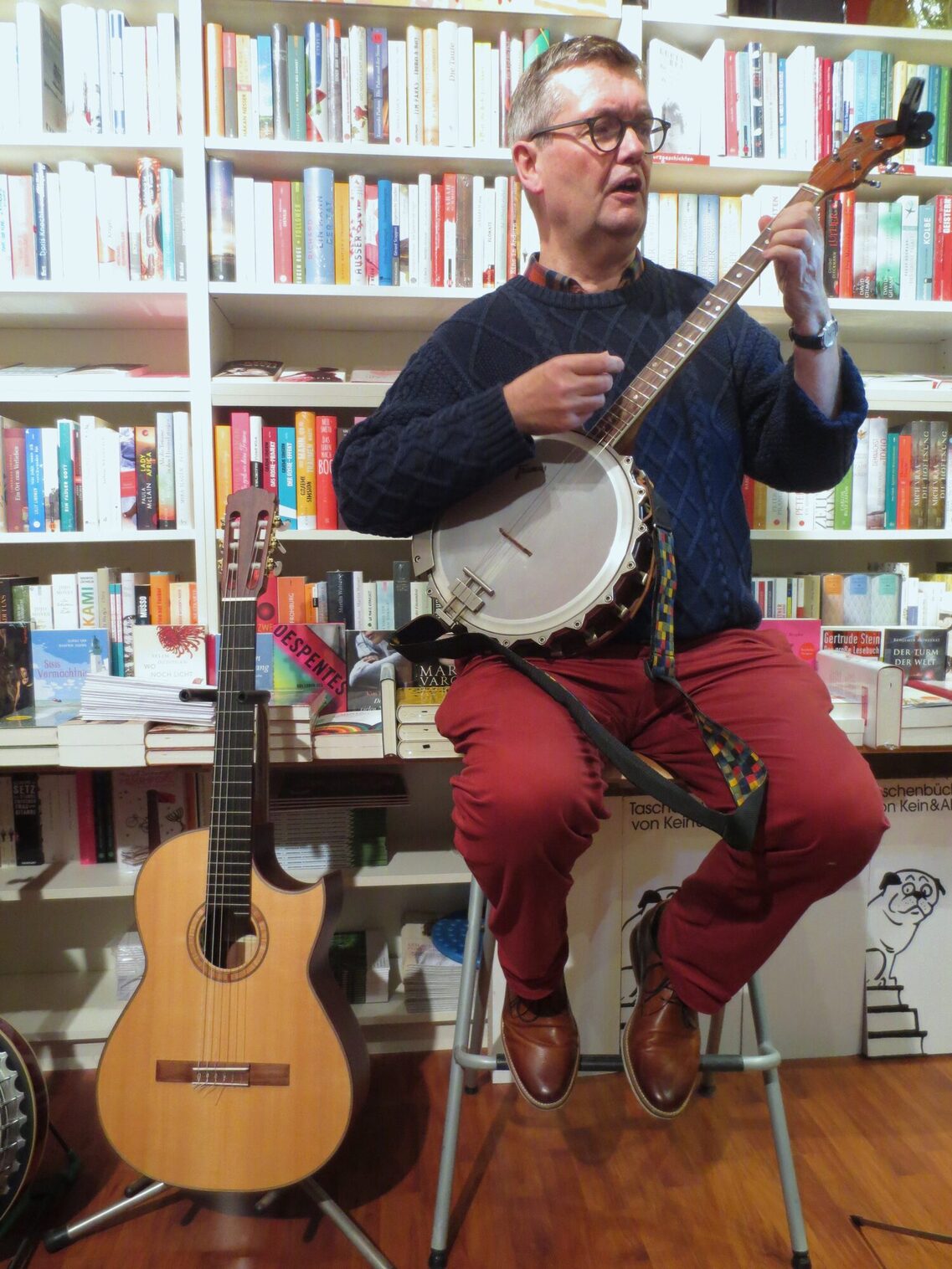 Ein Mann in blauem Pulli und bordeauxfarbener Hose sitzt auf einem Stuhl mit Perkussion vor einem Buchregal. Er wirkt sehr konzentriert.