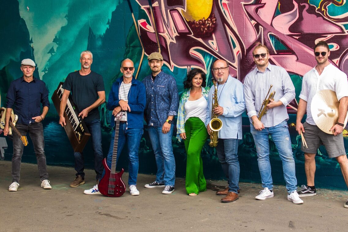 Renkli bir grafiti duvarının önünde ellerinde müzik aletleri olan 8 kişi. Resim çok renkli ve pozitif.