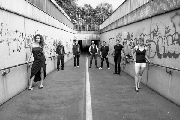 Gruppenfoto am Übergang zu einer Unterführung, alle sieben Bandmitglieder stehen rechts und links versetzt hintereinander. Das Bild ist in schwarz weiß. An der Wand der Unterführung sind Grafitti.