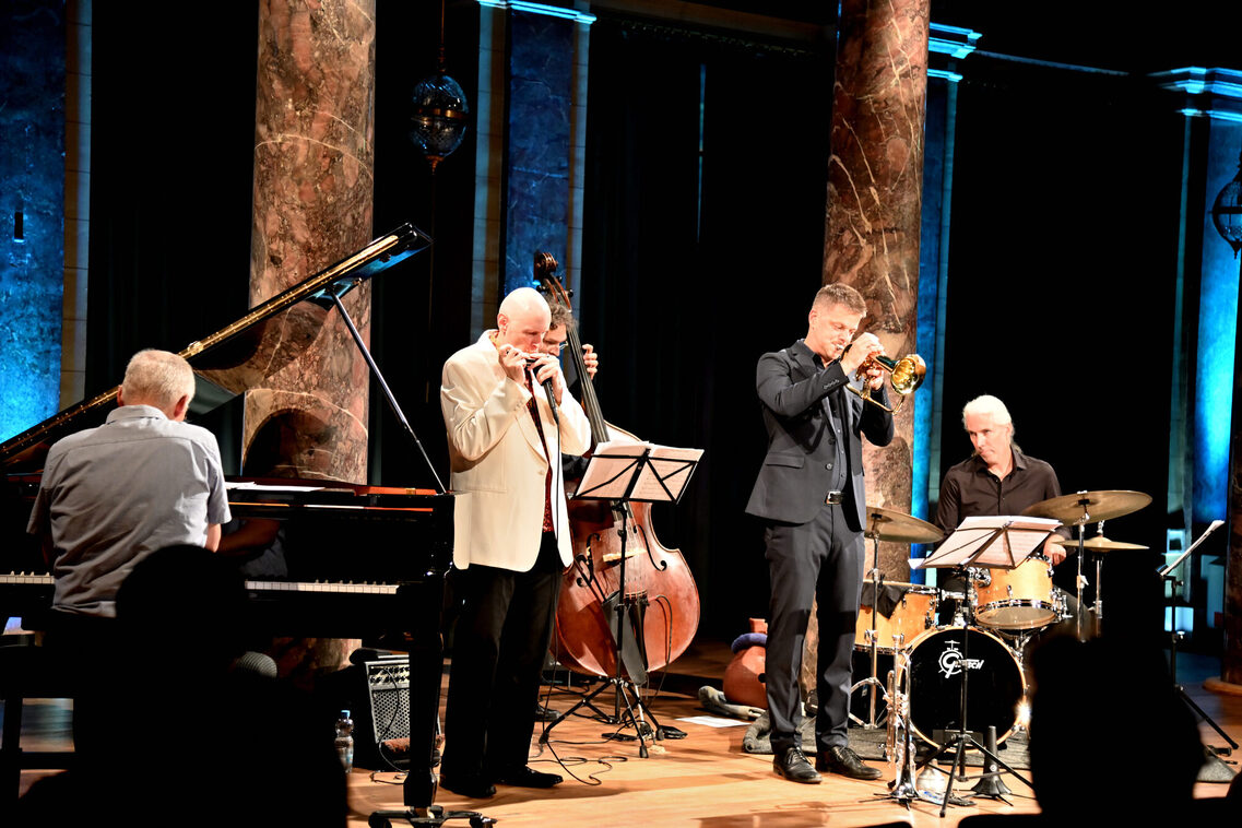 Вляво е мъж, седнал на роял, в средата отпред вляво е мъж с хармоника, в средата отзад е мъж на бас китара, в средата отпред вдясно е мъж на флюгелхорн, а вдясно е мъж на барабани.