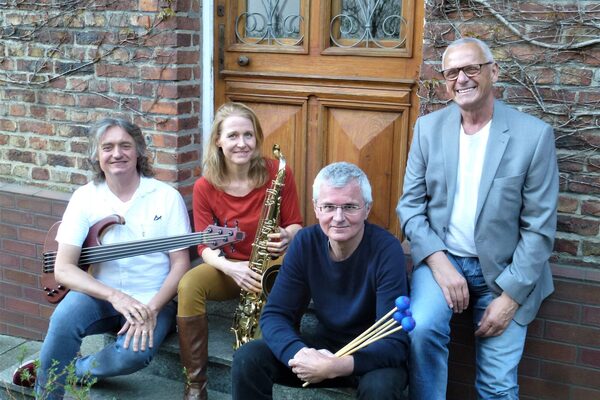 The band is sitting on a step in front of a front door. A man with an electric bass on the left, a woman with a saxophone in the middle left, a man with drumsticks in the middle right and a man leaning against the wall on the right.