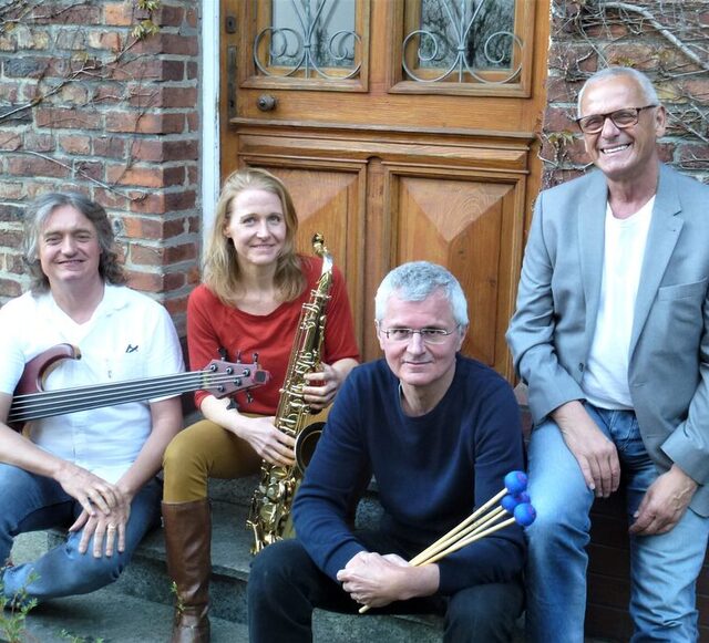 Die Band sitzt auf einer Stufe vor einer Haustür. Links ein Mann mit E-Bass, links in der Mitte eine Frau mit Saxofon, rechts in der Mitte ein Mann mit Drumsticks und rechts ein Mann an der Wand gelehnt.