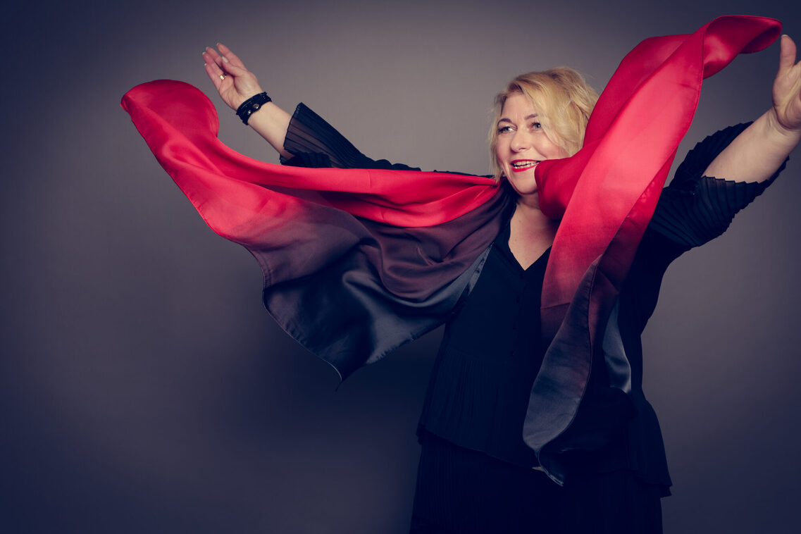 A blonde woman in black, throwing the red and black scarf around her neck upwards with her hands on either side.