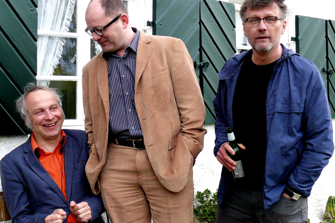 Στα αριστερά ένας άνδρας με μπλε κοστούμι και κόκκινο πουκάμισο χαμογελαστός και καθισμένος σε ένα παγκάκι, στο κέντρο ένας άνδρας με καφέ σακάκι και γυαλιά όρθιος, στα δεξιά ένας άνδρας με μπλε πουκάμισο και γυαλιά με μια μπύρα στο χέρι. Μόνο αυτός κοιτάζει στην κάμερα και οι άλλοι δύο φαίνεται να κάνουν ο ένας τον άλλον να γελάει.