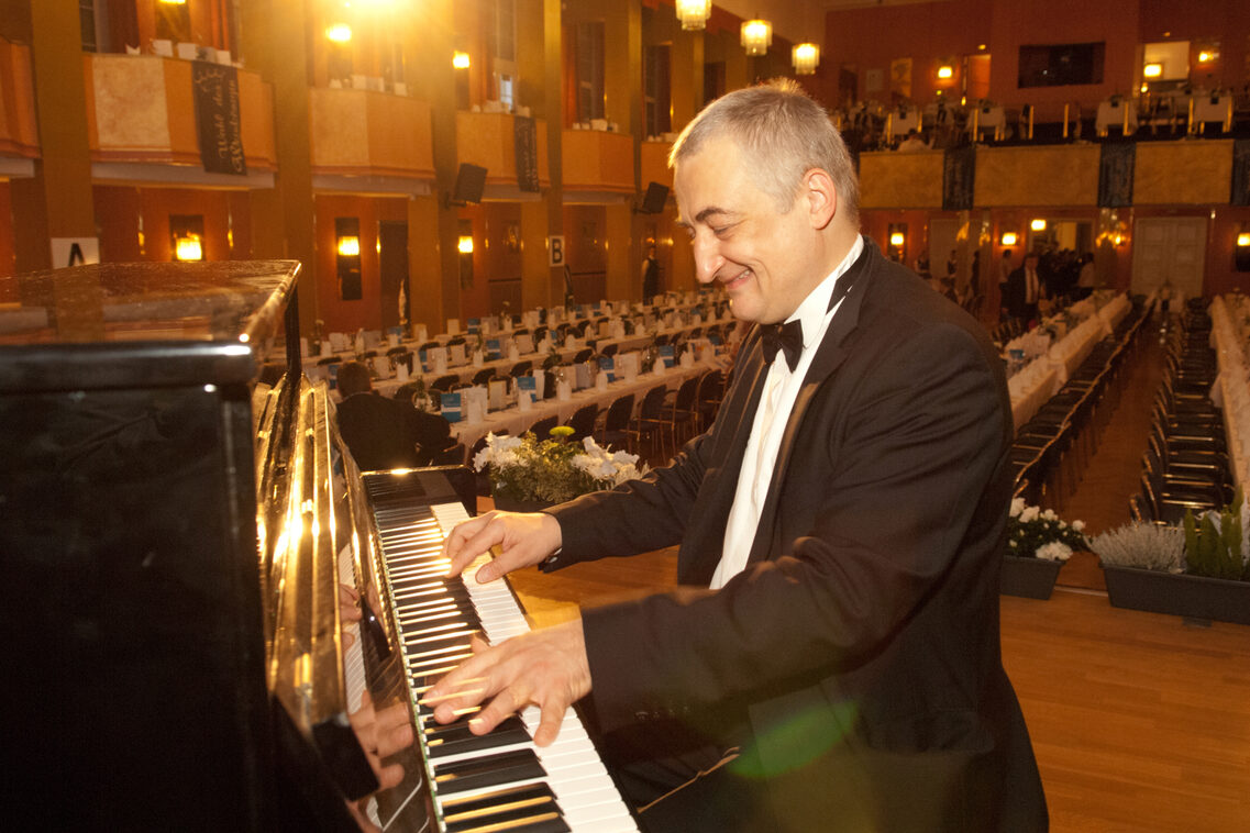 Mann in schwarzem Smoking am Piano in großem Saal. Alles ist in einem goldenen Licht.
