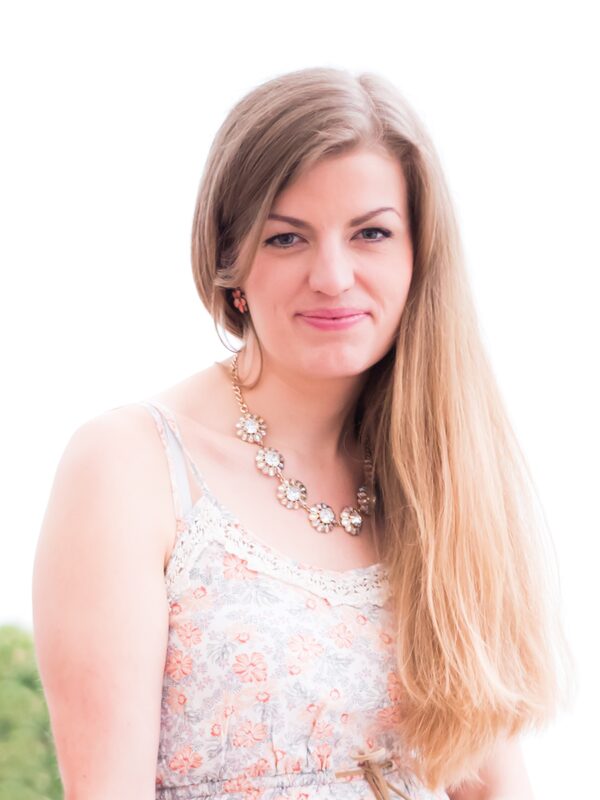 Eine blonde Frau in geblümten Kleid und mit großer Kette. Das Bild insgesamt ist sehr hell und man sieht im Hintergrund kaum etwas. Die Künstlerin lächelt in die Kamera.
