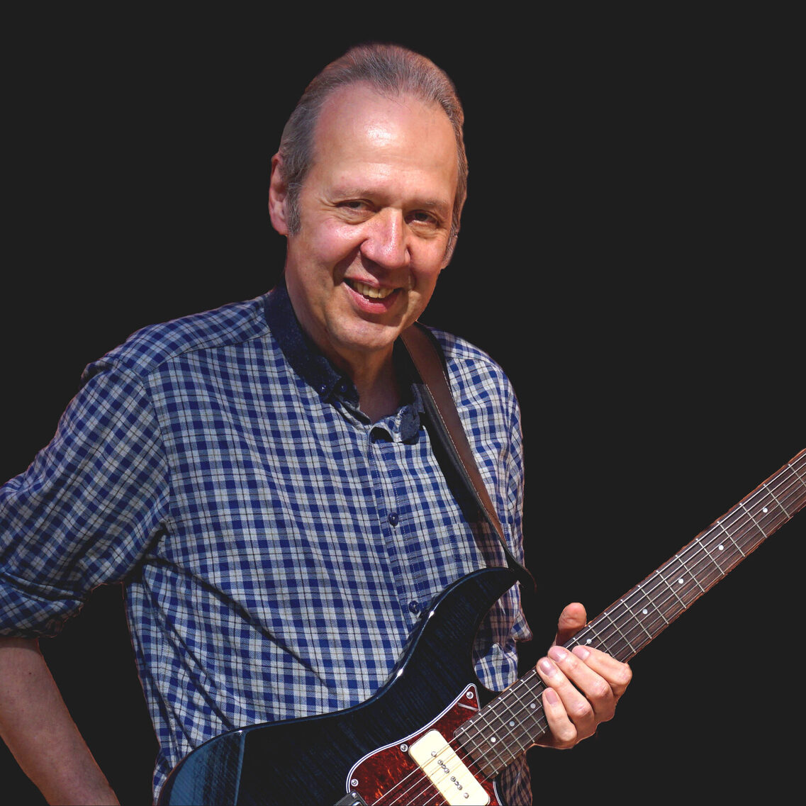 رجل يرتدي قميصاً أزرق وأبيض مزركشاً وفي يده جيتار كهربائي. الخلفية سوداء بالكامل والتقطت الصورة في استوديو تصوير.