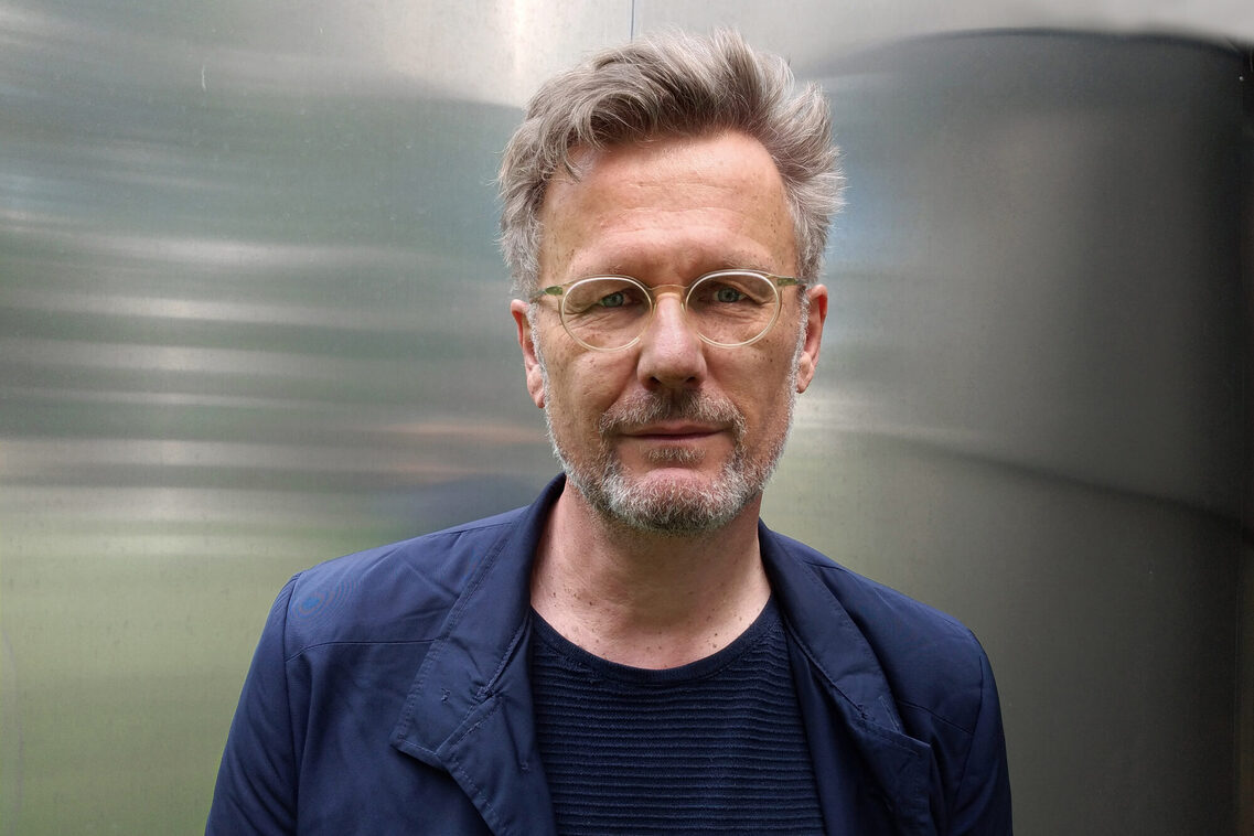 Frontalfoto, Mann mit kurzen grauen Haaren in dunkelblauer Jacke mit Brille. Der Hintergrund ist silber und spiegelt etwas, wird dadurch aber verschwommen und Uwe Oberg tritt mehr in den Vordergrund.