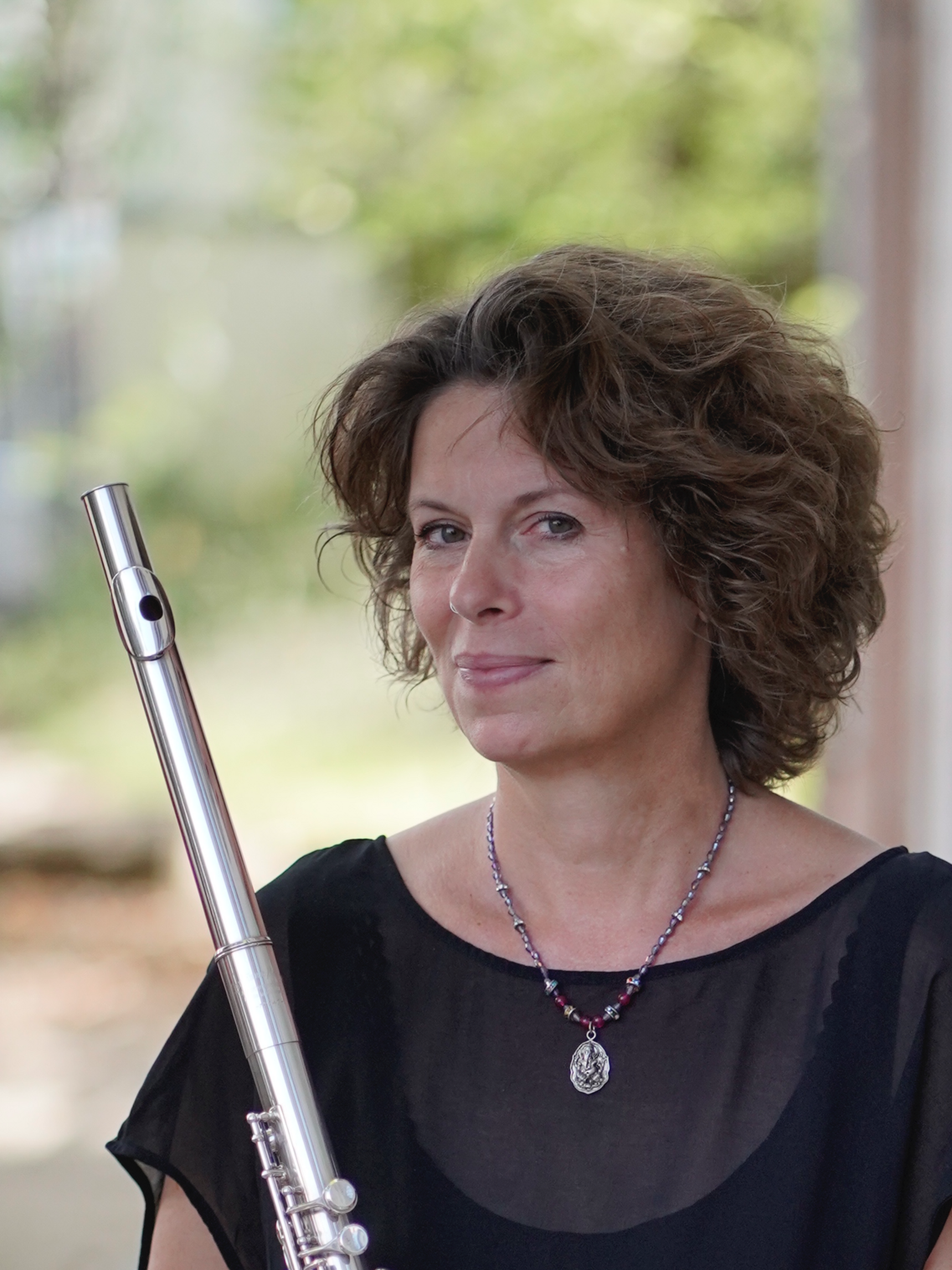 Frau mit dunklen kurzen Haaren und Querflöte in der Hand. Sie guckt seitlich in die Kamera und hat eine schwarze Bluse an.