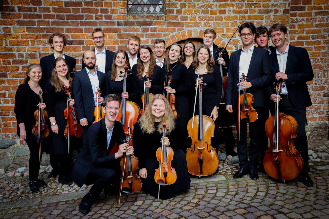 Das Ensemble steht zusammen mit ihren Instrumenten vor einer Wand. Die Besetzung besteht aus Cellos, Bratschen und Geigen.