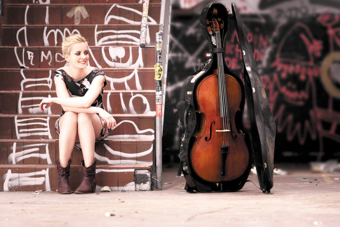 Mara sitzt links auf einer Treppe, die mit Graffiti bedeckt ist. Rechts neben ihr steht ein Cello und Mara guckt dort hin.