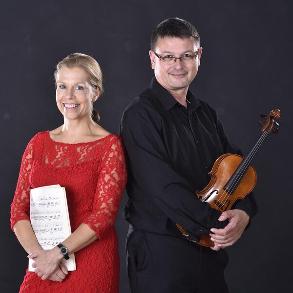 Links steht eine Frau im roten Kleid mit Musiknoten in der Hand und rechts ein Mann mit einer Violine.