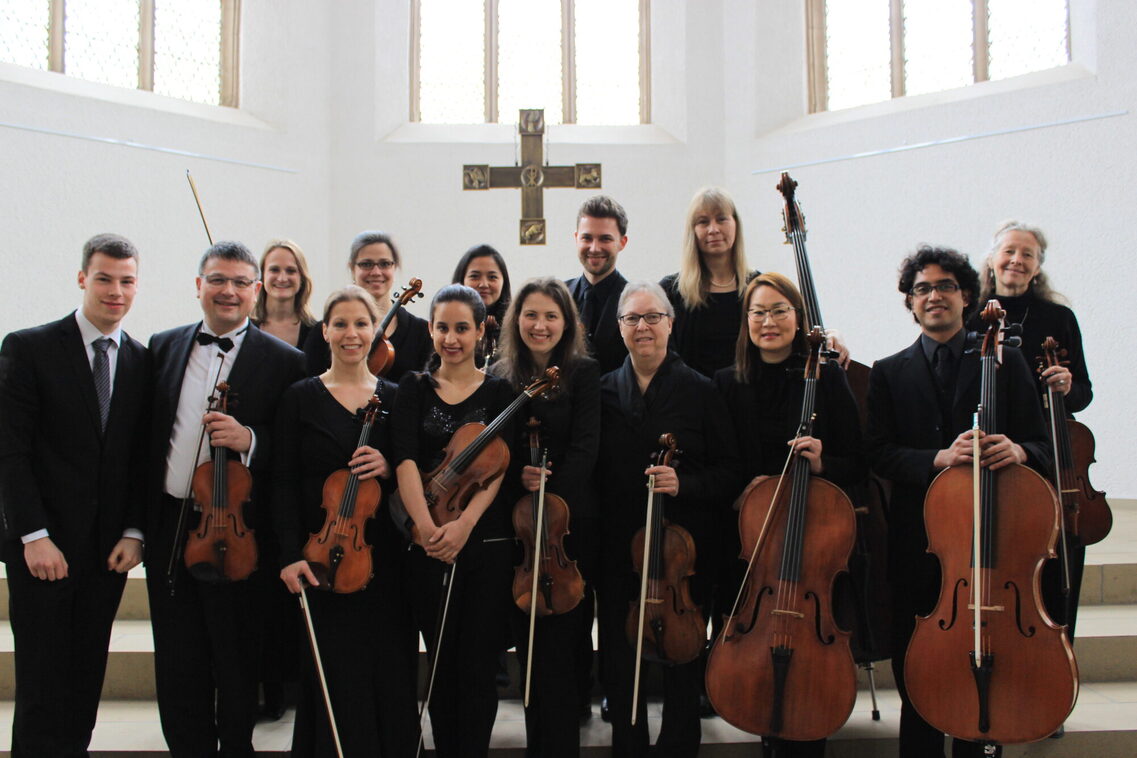 Es stehen viele Menschen mit ihren Instrumenten in der Kirche. Es sind alles Streichinstrumente und alle Mitglieder lächeln in die Kamera.