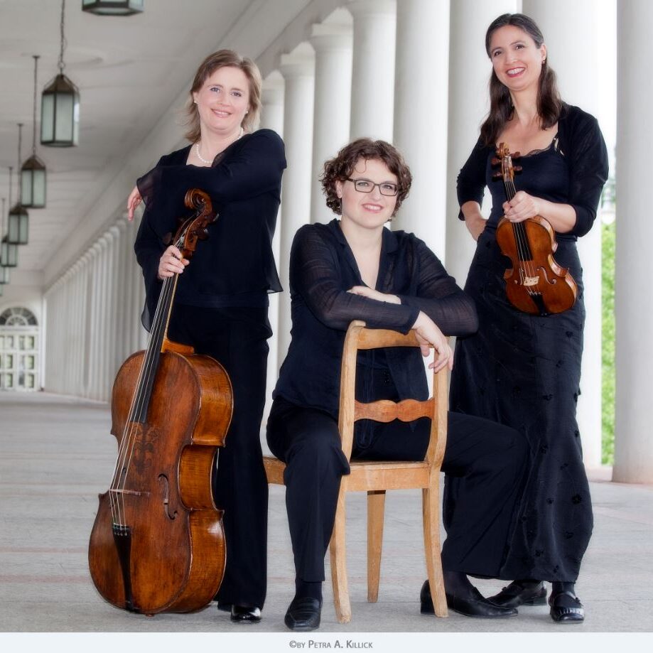 Tres mujeres vestidas de negro en la columnata del teatro de Wiesbaden. La mujer de la izquierda sostiene un violonchelo, la del centro está sentada en una silla de espaldas y la de la derecha sostiene un violín. Las tres mujeres sonríen.