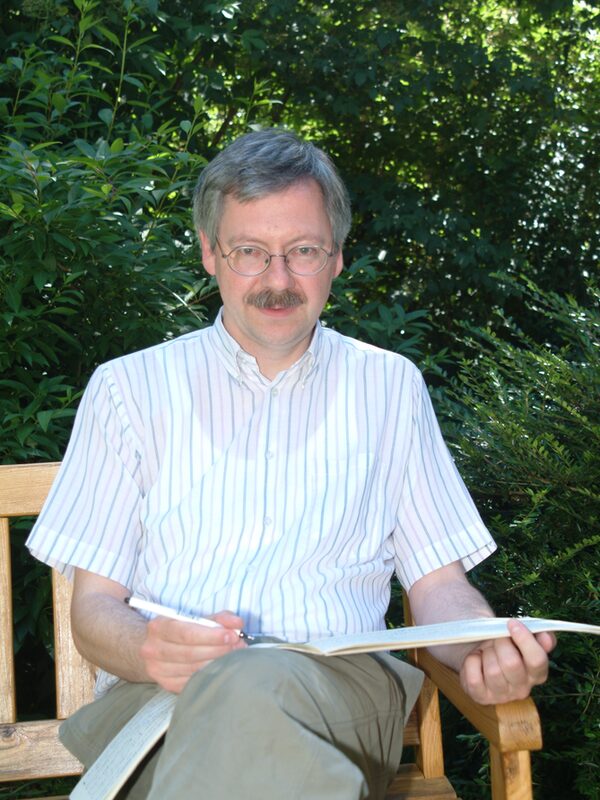 Burkhard Mohr sitzt auf einer Bank in einem Garten und auf seinen Beinen liegt ein Heft, in welches er rein schreibt. Er guckt in die Kamera.