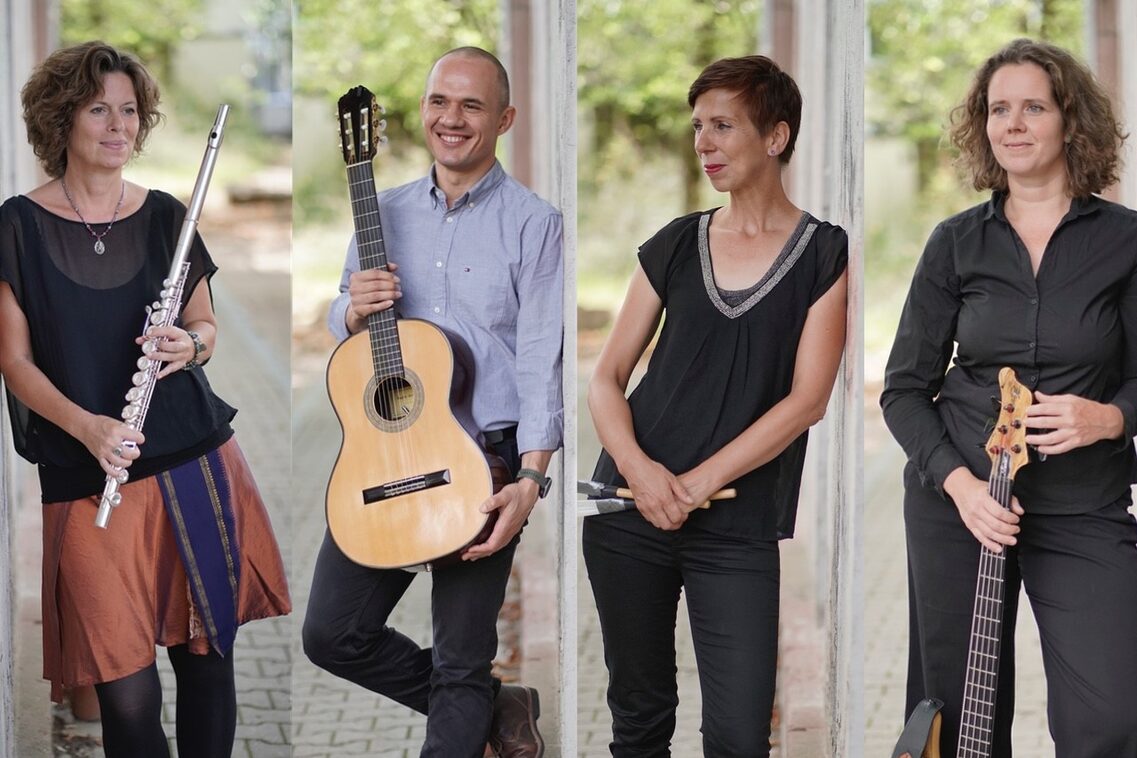 صورة مجمعة للفرقة. امرأة مع ناي على اليسار، ورجل مع جيتار في المنتصف الأيسر، وامرأة مع إيقاع في المنتصف الأيمن وامرأة مع إيقاع في المنتصف الأيمن وامرأة مع جيتار على اليمين.