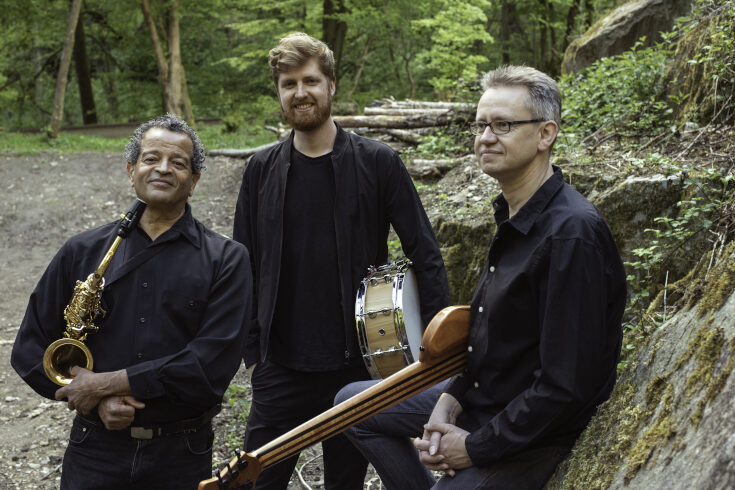 El trío JJJ de pie sobre las piedras con ropa negra. Todos sostienen sus instrumentos.