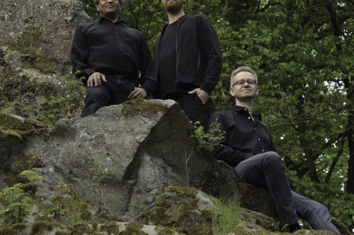 Das JJJ-Trio sitzend auf Steinen in schwarzer Kleidung. Alle schauen runter zu der Kamera.
