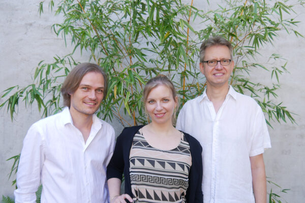 Il trio Jazzil è in piedi con i propri strumenti. Si trovano di fronte a un muro bianco e a un piccolo albero. Tutti sorridono alla telecamera.