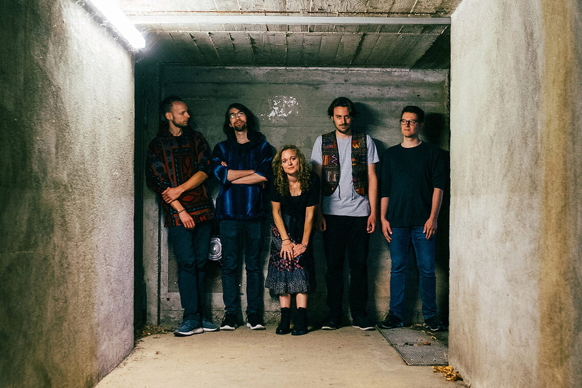 رجلان على اليسار، وامرأة في الوسط، ورجلان في القبو/الممر السفلي على اليمين. يقف الجميع بشكل عرضي جداً والمرأة منحنية.