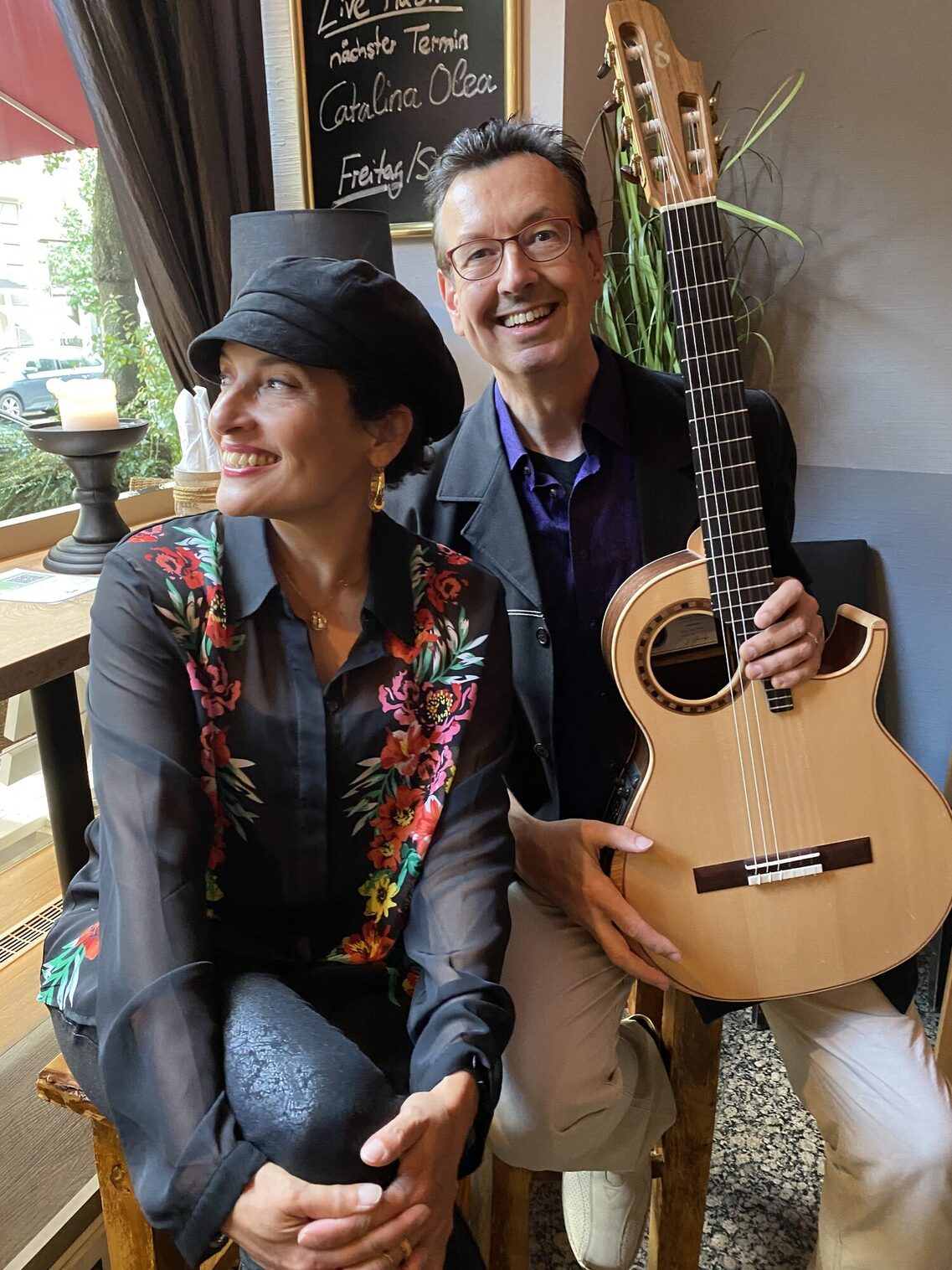 Seduta alla finestra, a sinistra, una donna con un cappellino nero a palloncino, con le gambe incrociate e lo sguardo rivolto verso la finestra, a destra un uomo con una giacca nera e una chitarra in grembo, che sorride all'obiettivo.