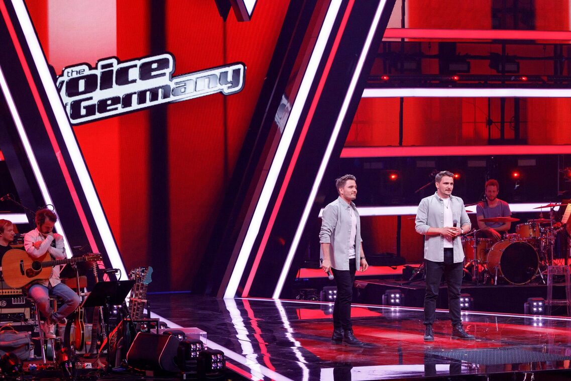 Kingtwiins auf der Bühne von "The Voice of Germany". Alles ist sehr rot und die Zwillinge sehen aufgeregt aus.