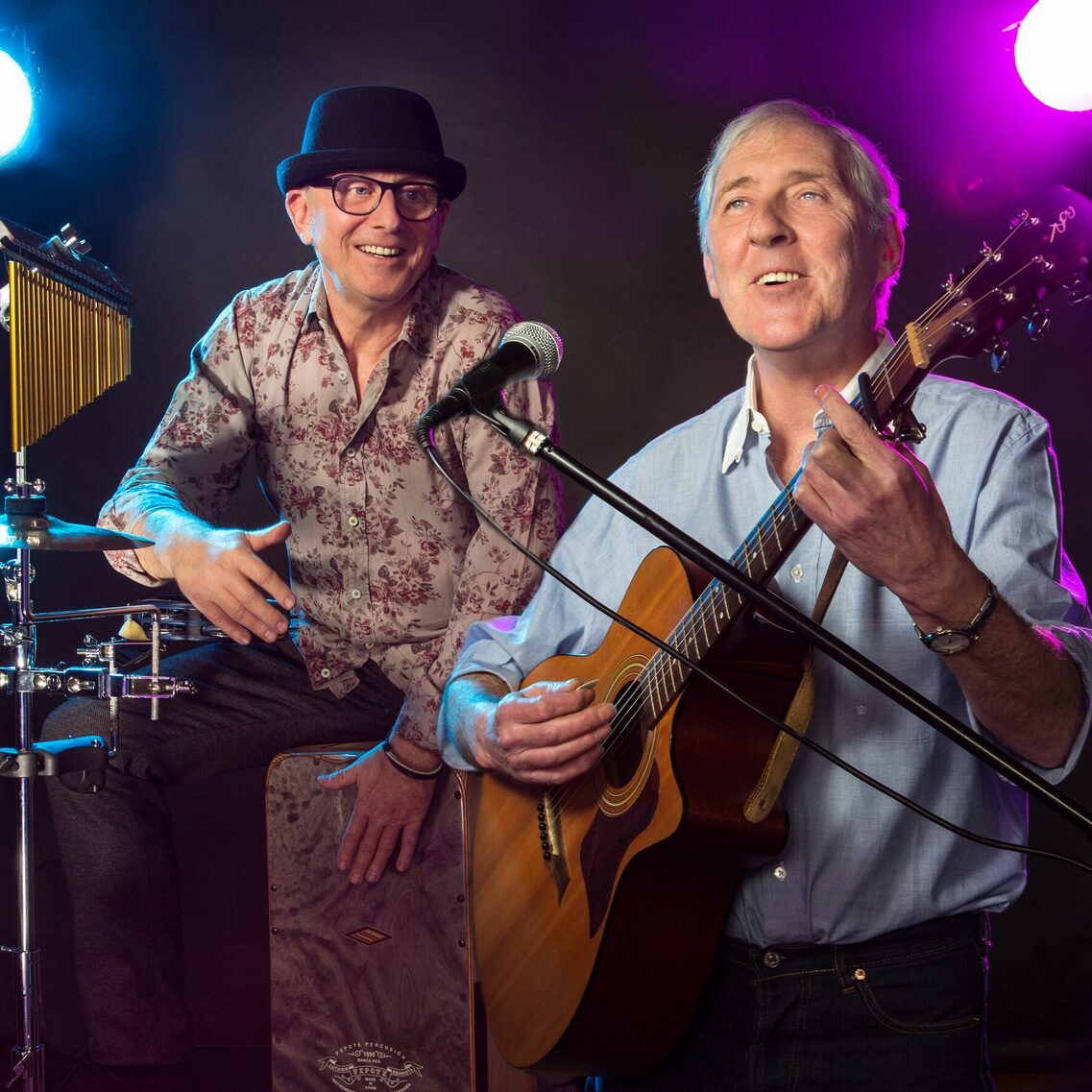 Отляво седи мъж с шапка на перкусиите, а отдясно - мъж с китара. Пред него има микрофон и изглежда, че той пее.