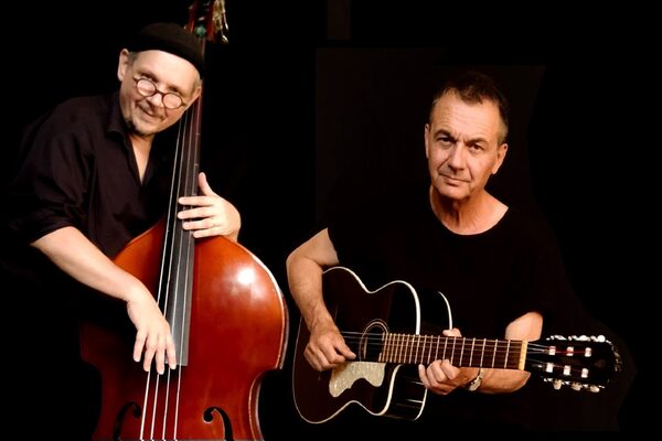 Links steht ein Mann mit einem Kontrabass und rechts neben ihm sitzt ein Mann mit einer Gitarre. Der Hintergrund ist schwarz und beide gucken in die Kamera.