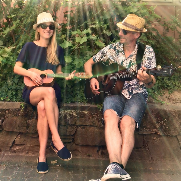 Links sitzt eine Frau mit einer Ukulele und rechts ein Mann mit einer Gitarre. Sonnenstrahlen, welche im Bild zu sehen sind, machen ein sommerliches Gefühl.
