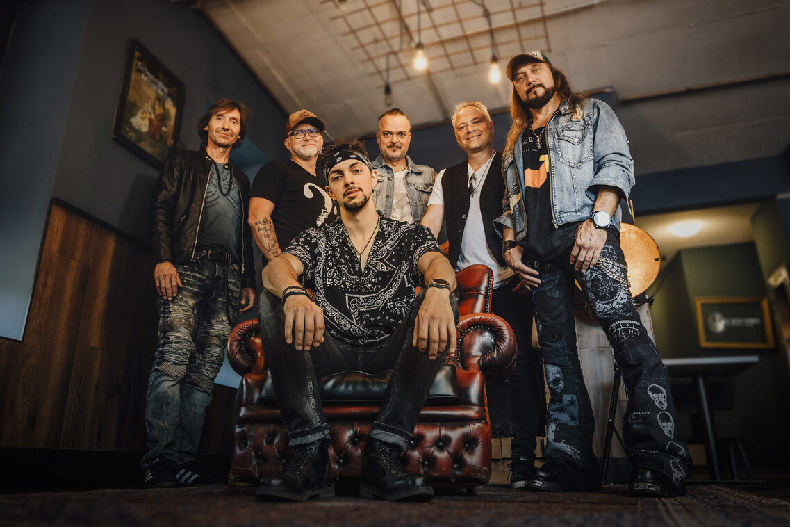 The band Juke Box Hero looks into the camera. One man is sitting on a chair and five are standing behind it. The photo was taken from below, so the band looks very confident and in a way powerful.