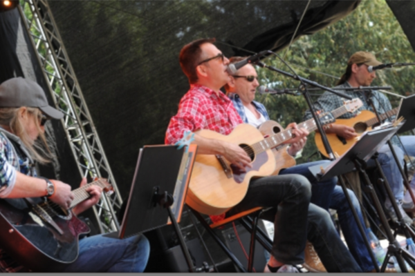 Quattro uomini, la band Acoustic Road, siedono sul palco e suonano tutti la chitarra. Uno di loro canta e sono tutti molto concentrati.