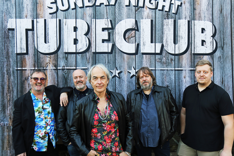 Der Sunday Night Tubeclub steht von einer Holzwand und lächelt lässig in die Kamera. Es sind fünf Männer und über Ihnen steht als Schriftzug der Name der Band.