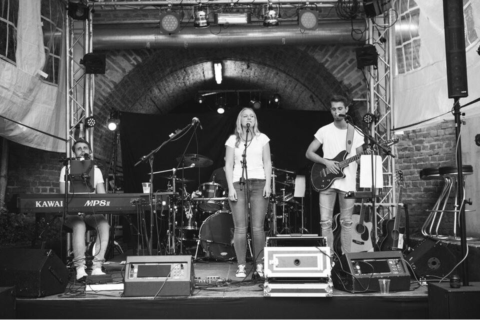 الخط الثالث على المسرح؛ رجل على البيانو، ورجل على الجيتار الكهربائي، وامرأة في الوسط أمام الميكروفون. الصورة بالأبيض والأسود.